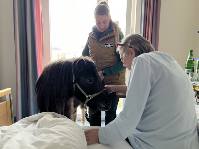 Shetlandpony Lena steht am Bett einer Bewohnerin. Im Hintergrund Ponybesitzerin Klara Dressler.