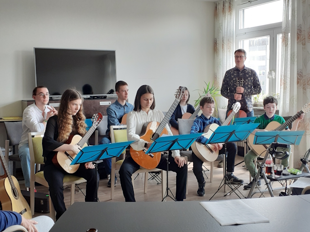Die Mitglieder des Gitarrenclubs sitzen auf Stühlen vor ihren Noten im Paul Marien Hospiz. Im Hintergrund steht der Leiter.